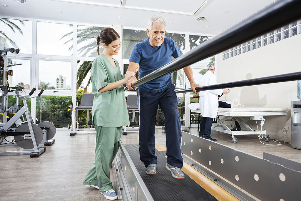 A Senior Man Walking Between Parallel Bars Being Helped By A Physical Therapist During Physical Therapy For Fall Prevention